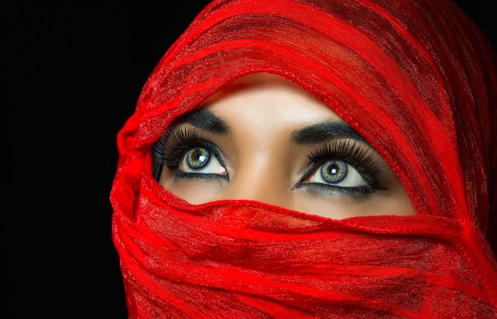 Bright blue eyes peer through a vibrant red silk scarf wrapped around a face against a dark background.