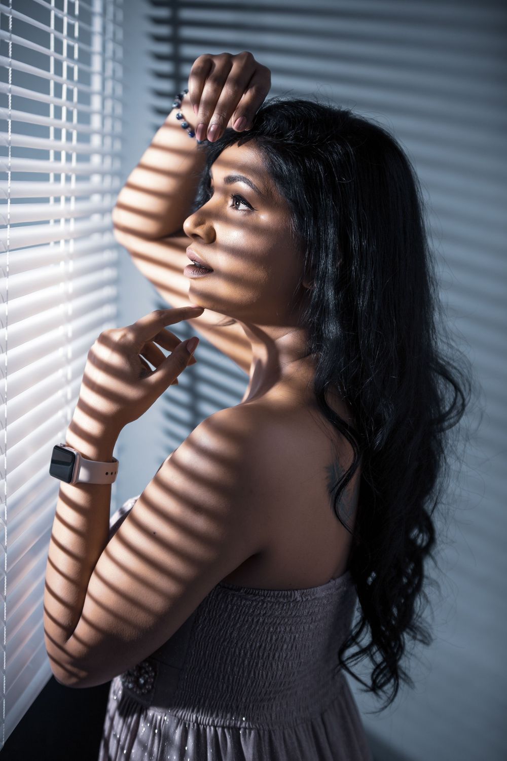 Dramatic portrait with sunlight casting venetian blind shadows across figure in grey dress near window.