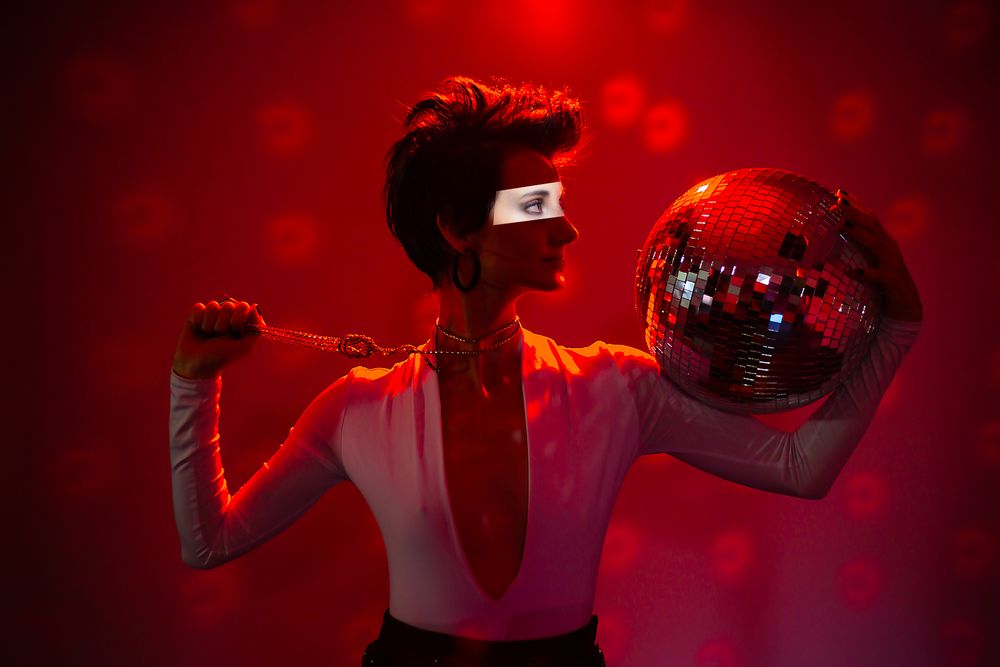 Disco-style figure in white shirt poses with glowing red disco ball against dramatic red background and lighting effects.