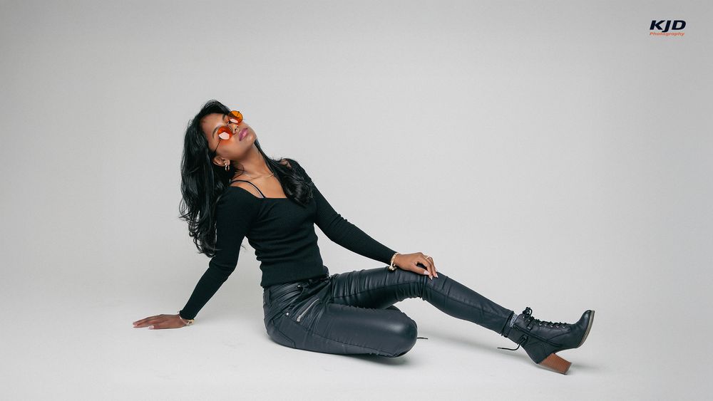 Woman in black leather jacket and pants poses elegantly on floor against white background.