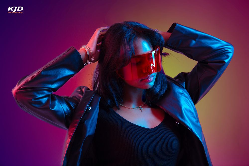 Woman in leather jacket and red sunglasses poses under vibrant neon purple and yellow studio lighting.