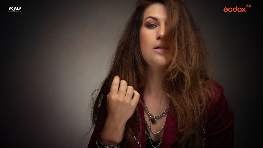 A person in a burgundy top with long tousled brown hair poses dramatically against a dark background.