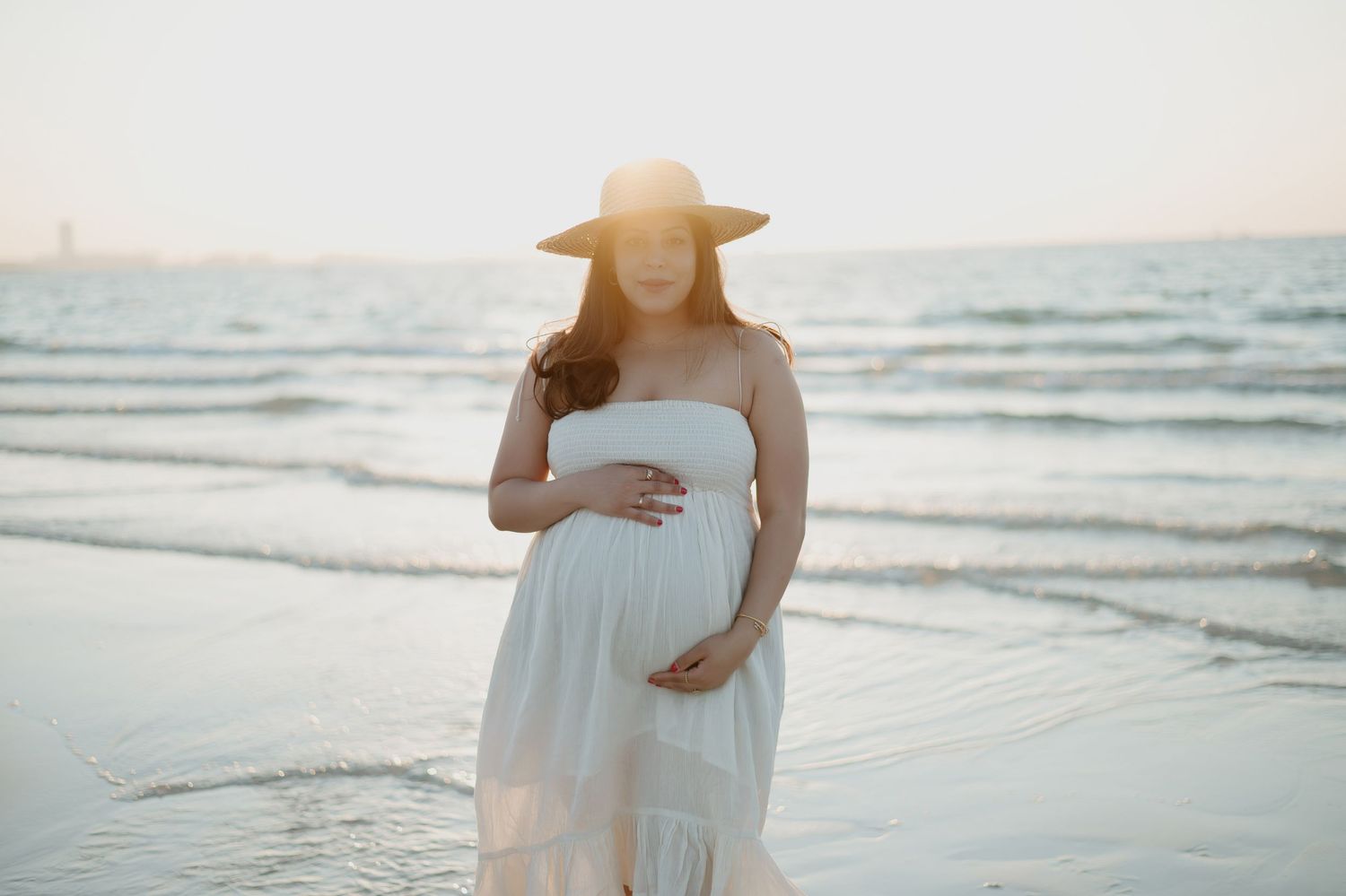 Pregnant woman in flowing white dress and sunhat poses on beach at golden hour.