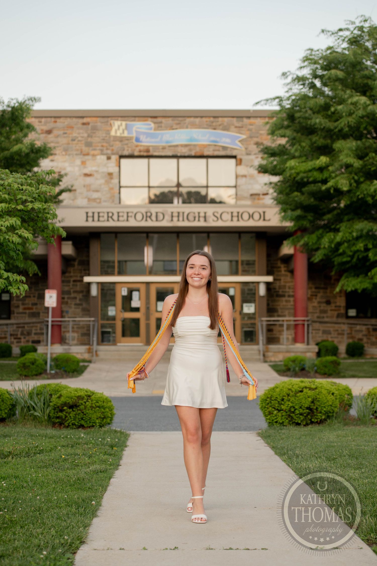 Graduate poses in front of Hereford High School entrance wearing a white dress on a sunny summer day.