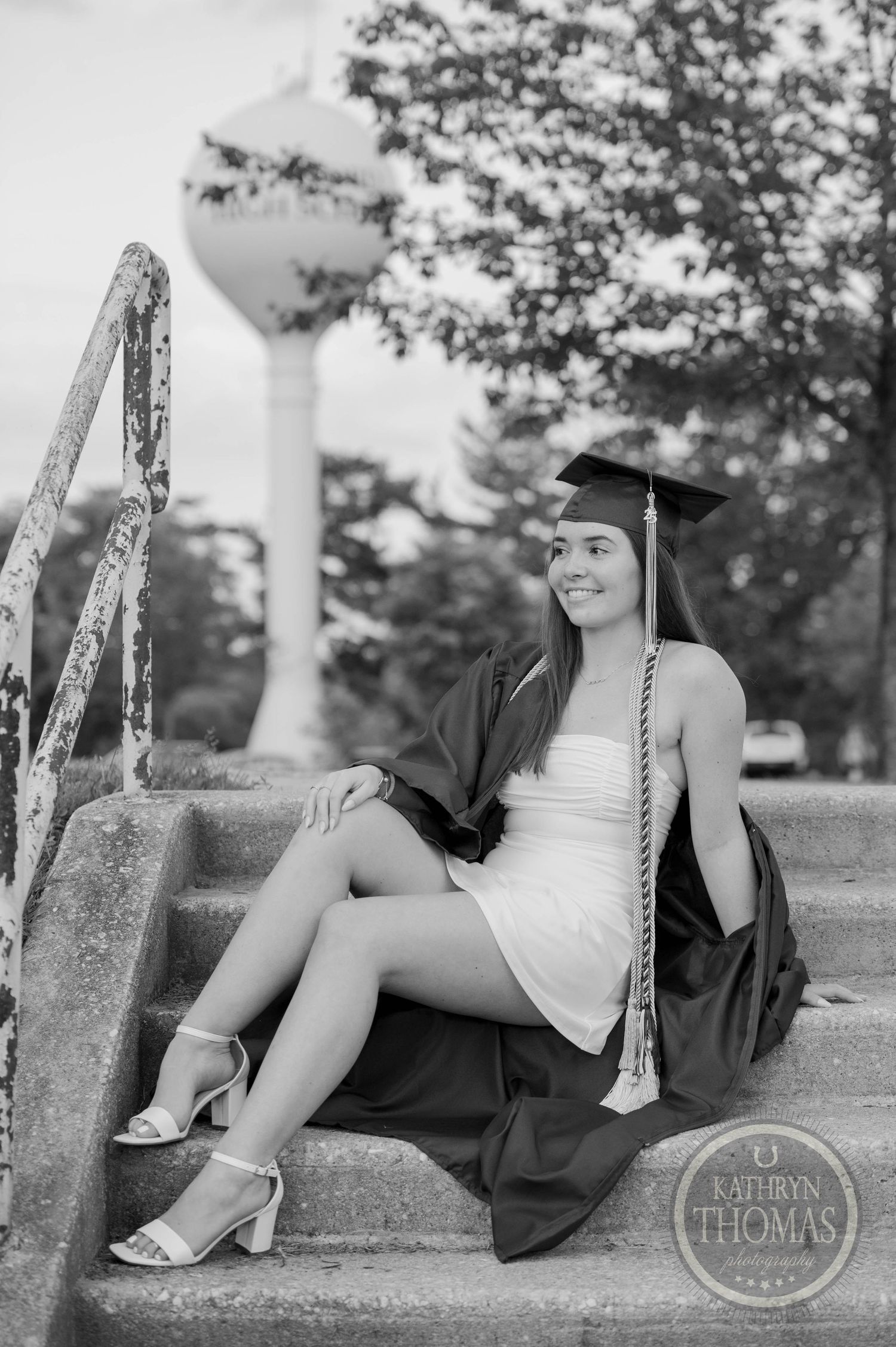 A graduate in maroon cap and gown sits on concrete steps with a water tower visible in the background.