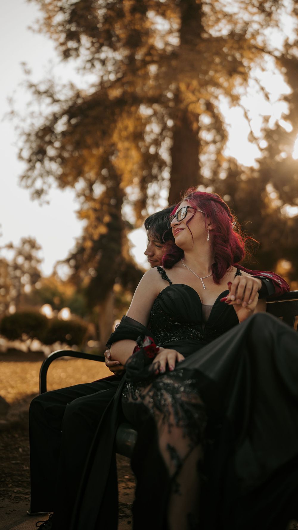 A person in a black dress relaxes on a bench surrounded by tall trees at sunset.