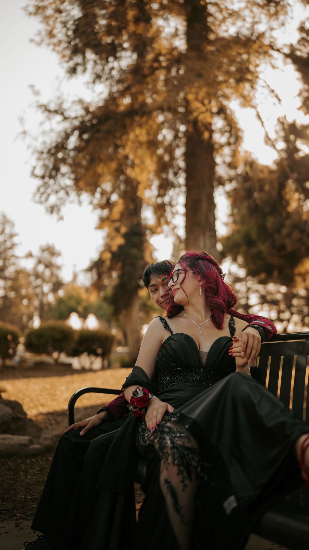 A gothic-styled figure in a black dress sits on a park bench at sunset with warm autumn lighting in the background.