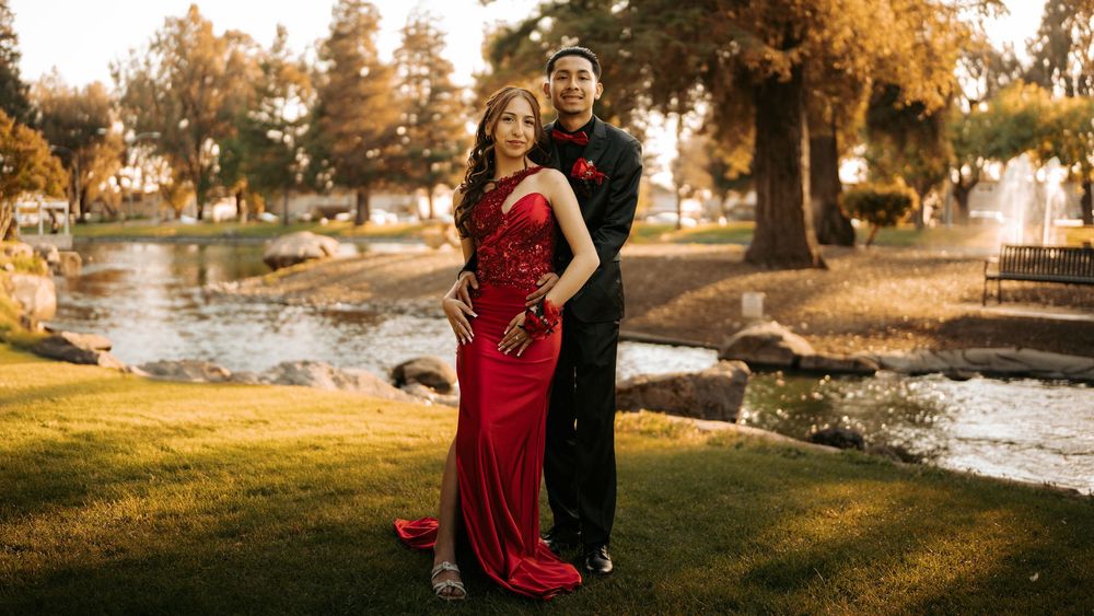 Couple in formal attire embraces by a pond at golden hour in a scenic outdoor park setting.