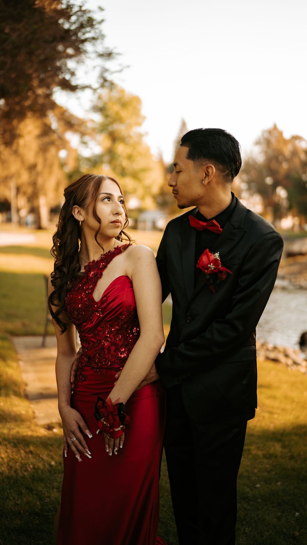 A formal couple in a red dress and black tuxedo pose romantically at sunset in a park setting.