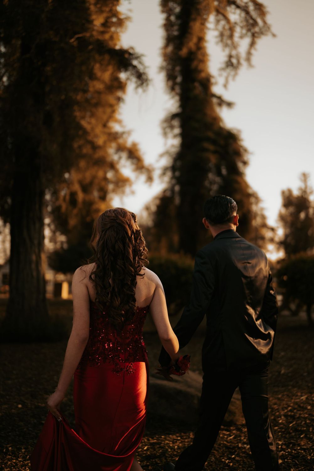 Couple in formal attire walking through trees at sunset with dramatic orange lighting.