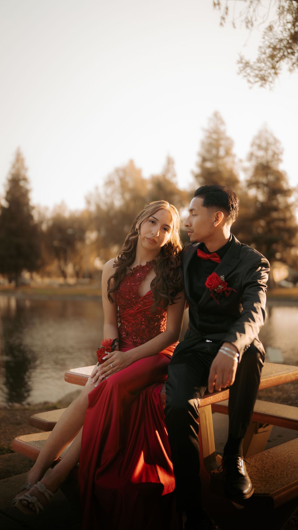 A couple in formal attire sits together on a dock by a lake at sunset, she in red and he in black with matching red details.