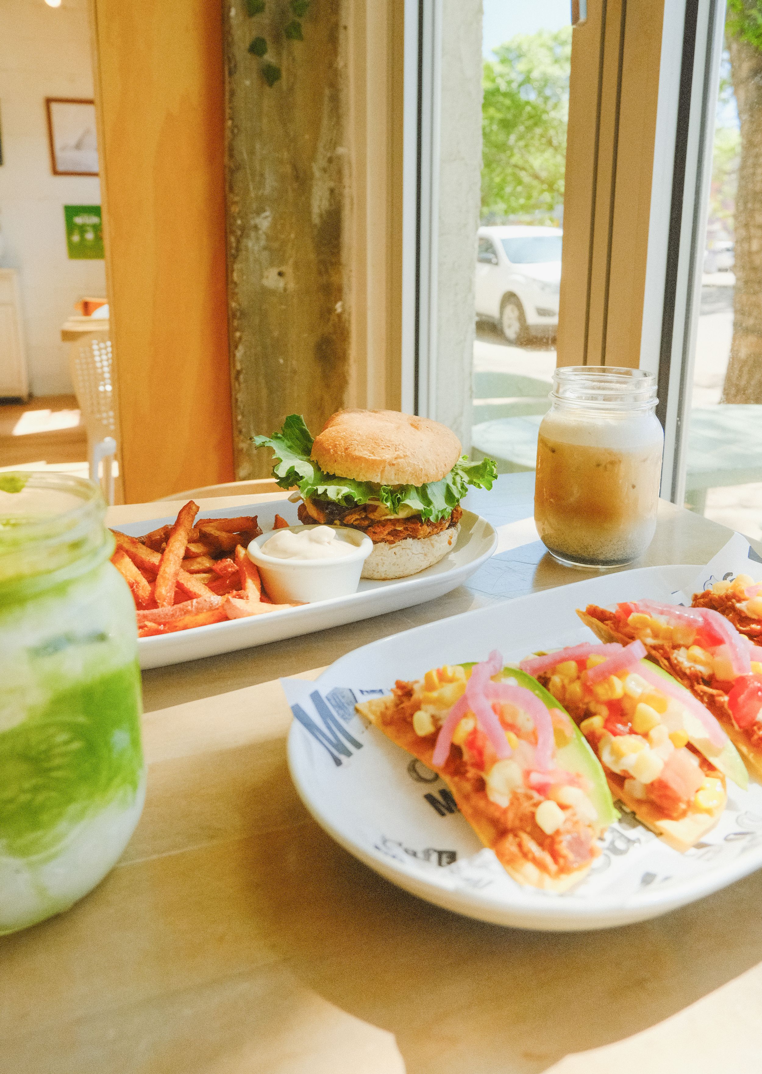 Artisanal burger and seafood dish served with french fries and colorful drinks on wooden table by sunny window.