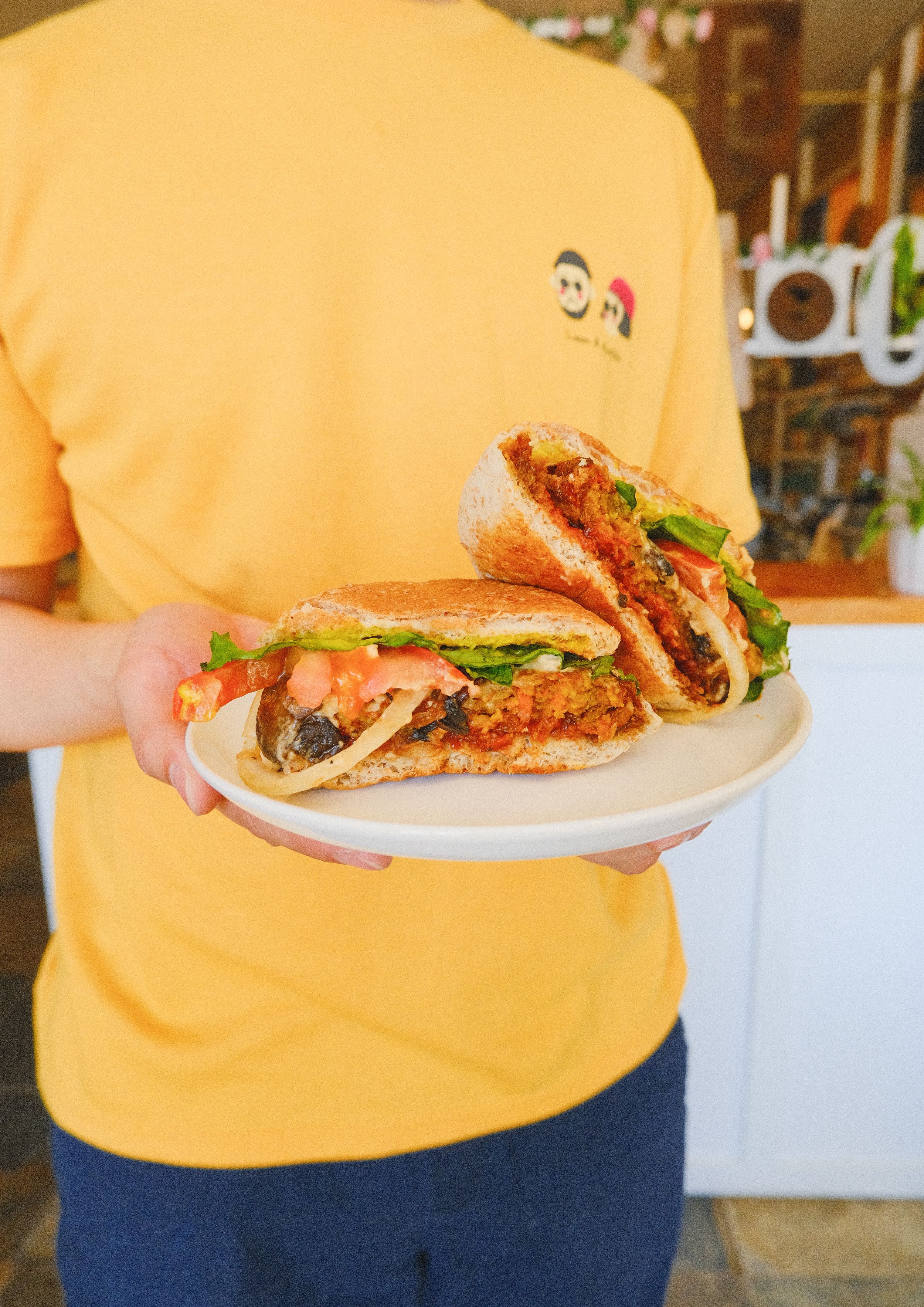 A person in a yellow shirt holds a plate with a sandwich or sub filled with meat and fresh vegetables.