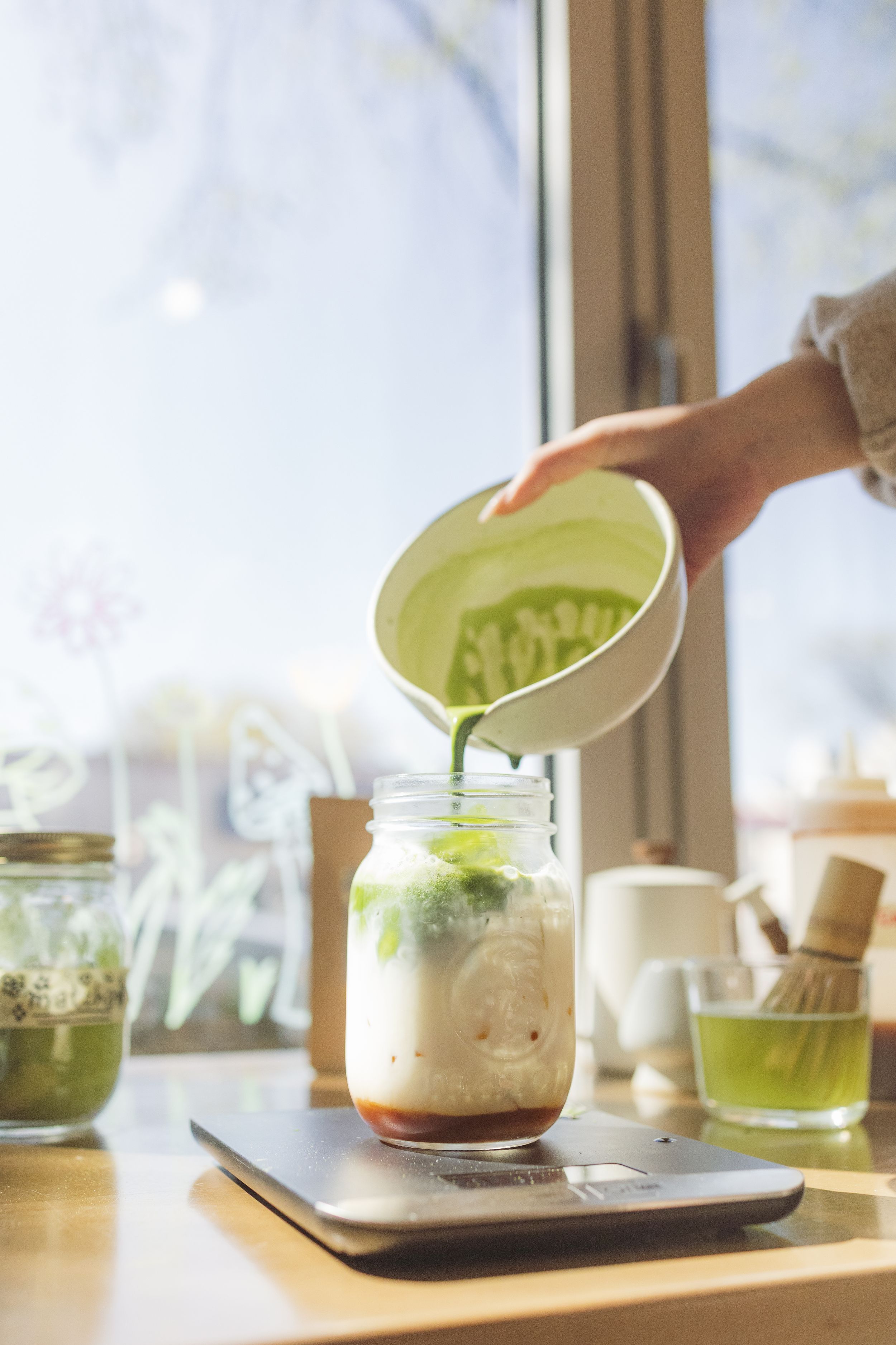 A series of four matcha green tea desserts in glass cups arranged in a row on a wooden surface near a window.