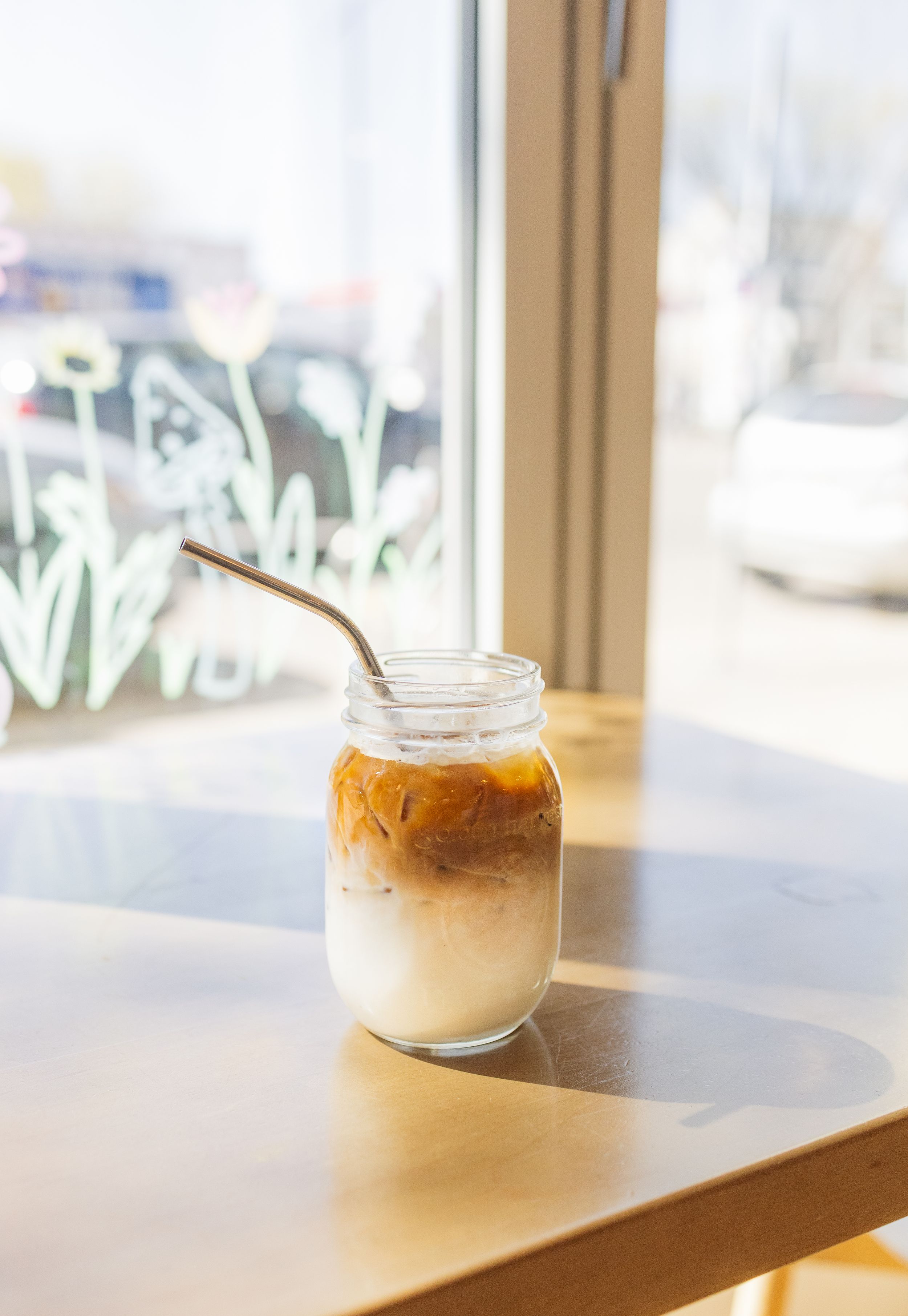 A glass jar of iced latte with layered milk and coffee sits by a window in soft natural lighting.