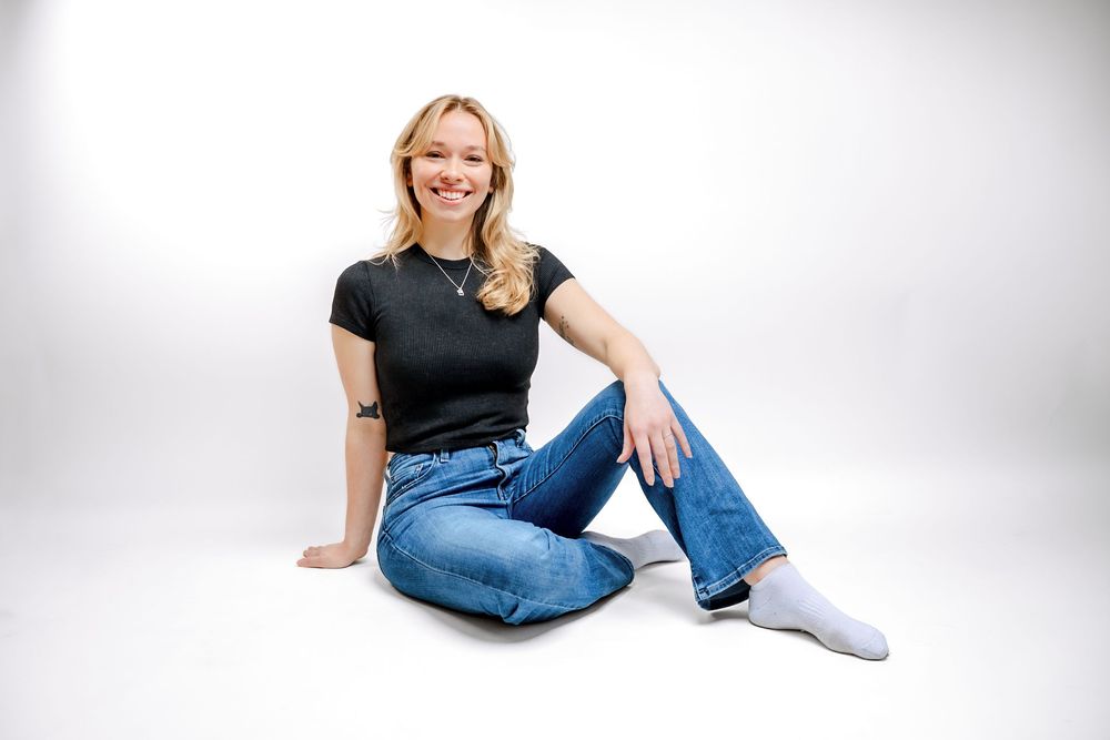 Young blonde woman smiling, seated on floor in black t-shirt, blue jeans, and white socks against white background.