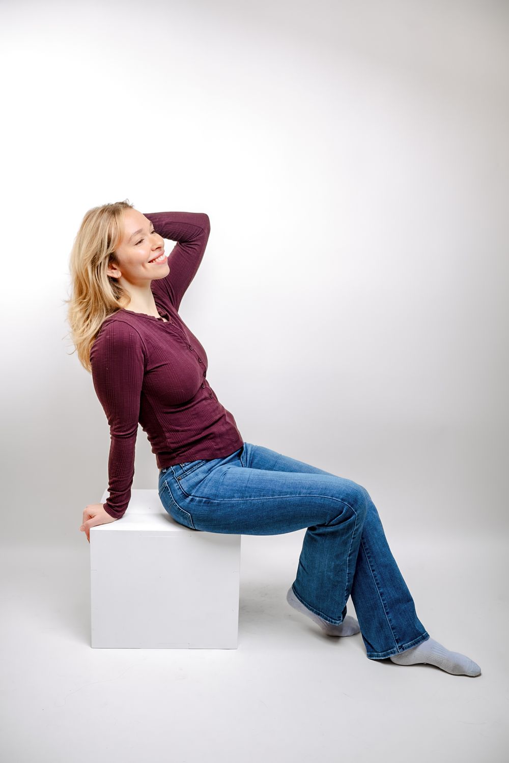 Smiling blonde woman sitting on a white cube, wearing burgundy top and flared jeans, posing in a studio.