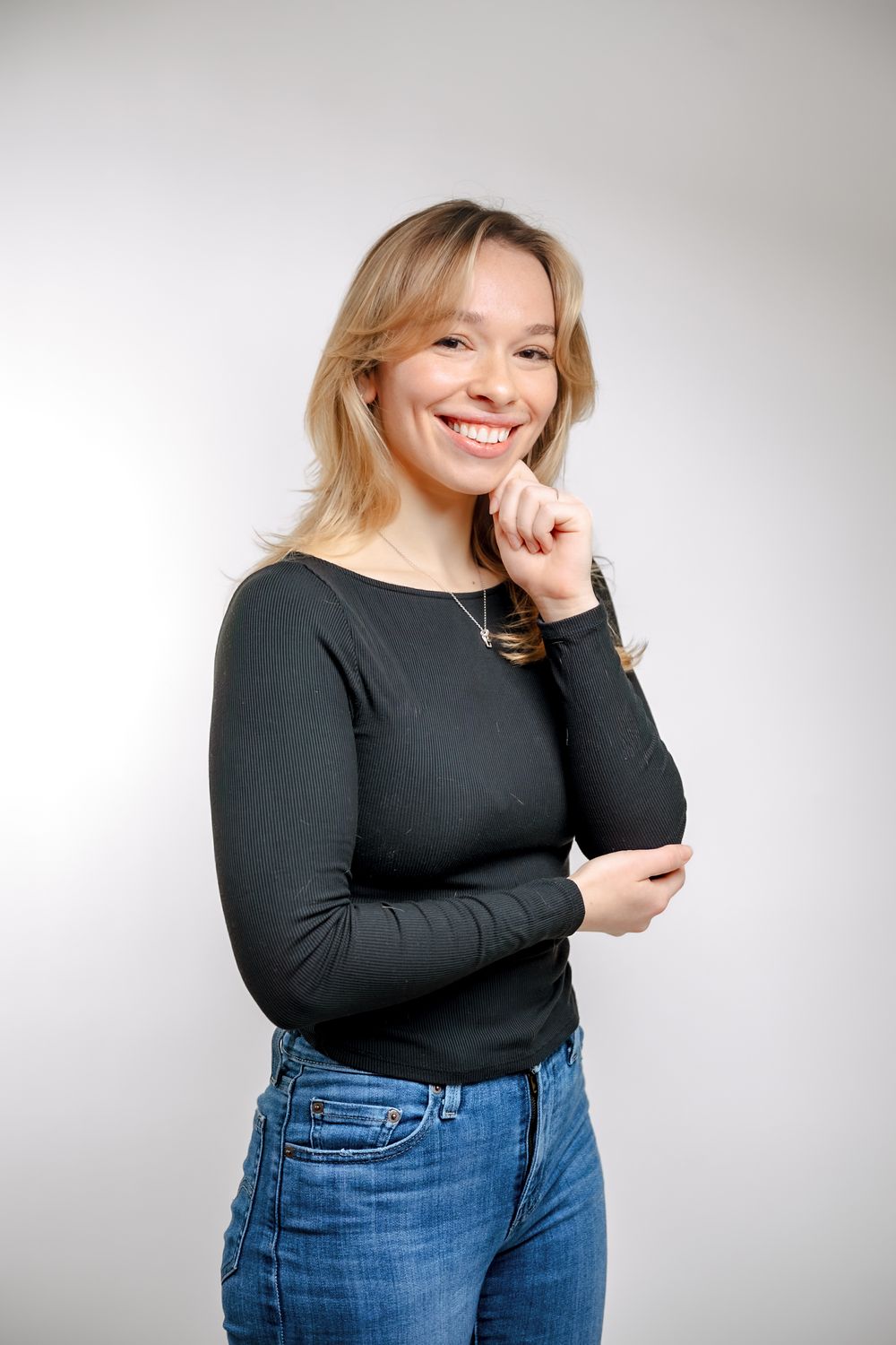 Smiling young woman with blonde hair wearing a black ribbed long-sleeve top and blue jeans posing against a white background.