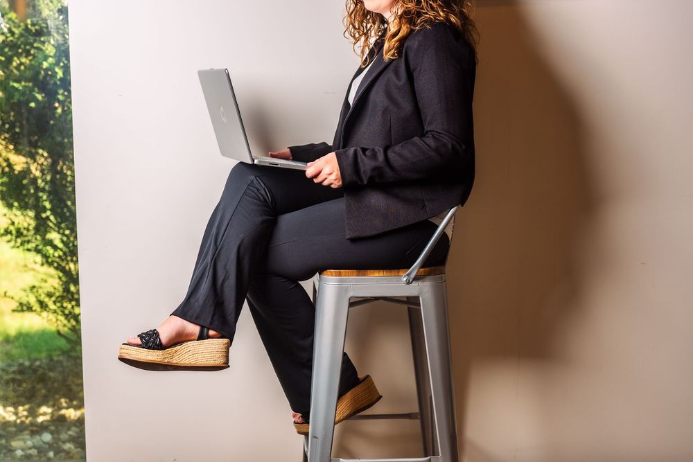 Woman working on laptop while sitting on modern metal stool in office space.