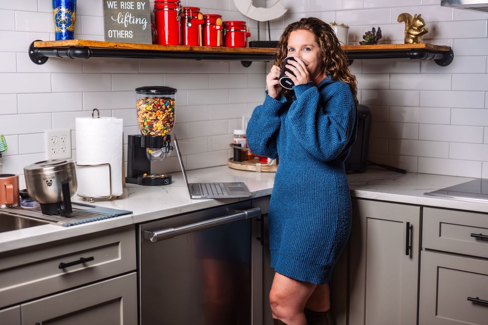 Woman in blue sweater dress enjoying coffee in modern kitchen with white tile backsplash.