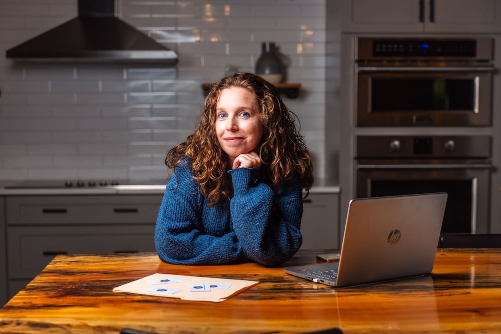 Person in blue sweater working at wooden table with laptop and papers in modern kitchen setting.