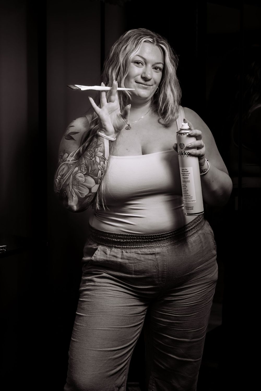 Person holding hair care product bottles while posing in black and white salon portrait.