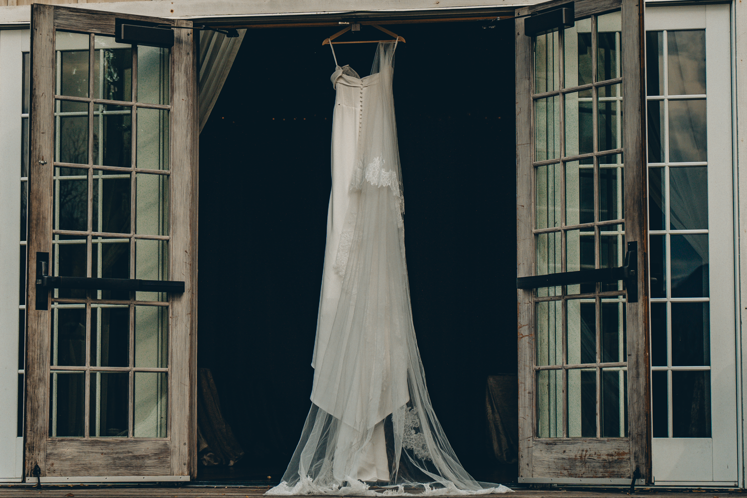 Elegant white wedding dress hanging in window frame, with delicate lace details against dark background.