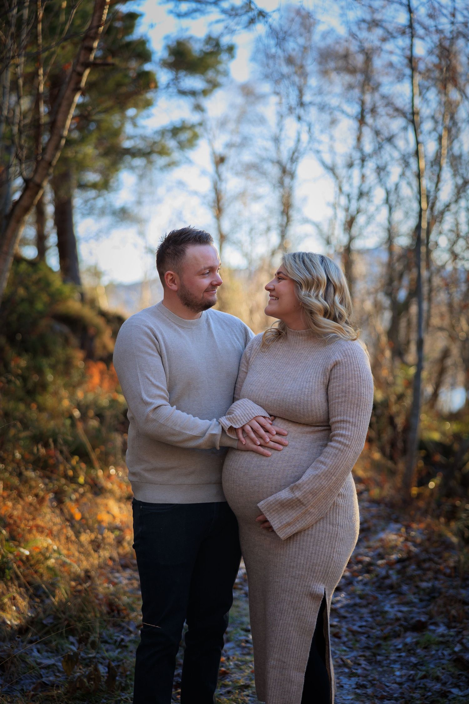 A couple in cozy sweaters share an intimate moment outdoors during autumn, with the expectant mother cradling her baby bump.