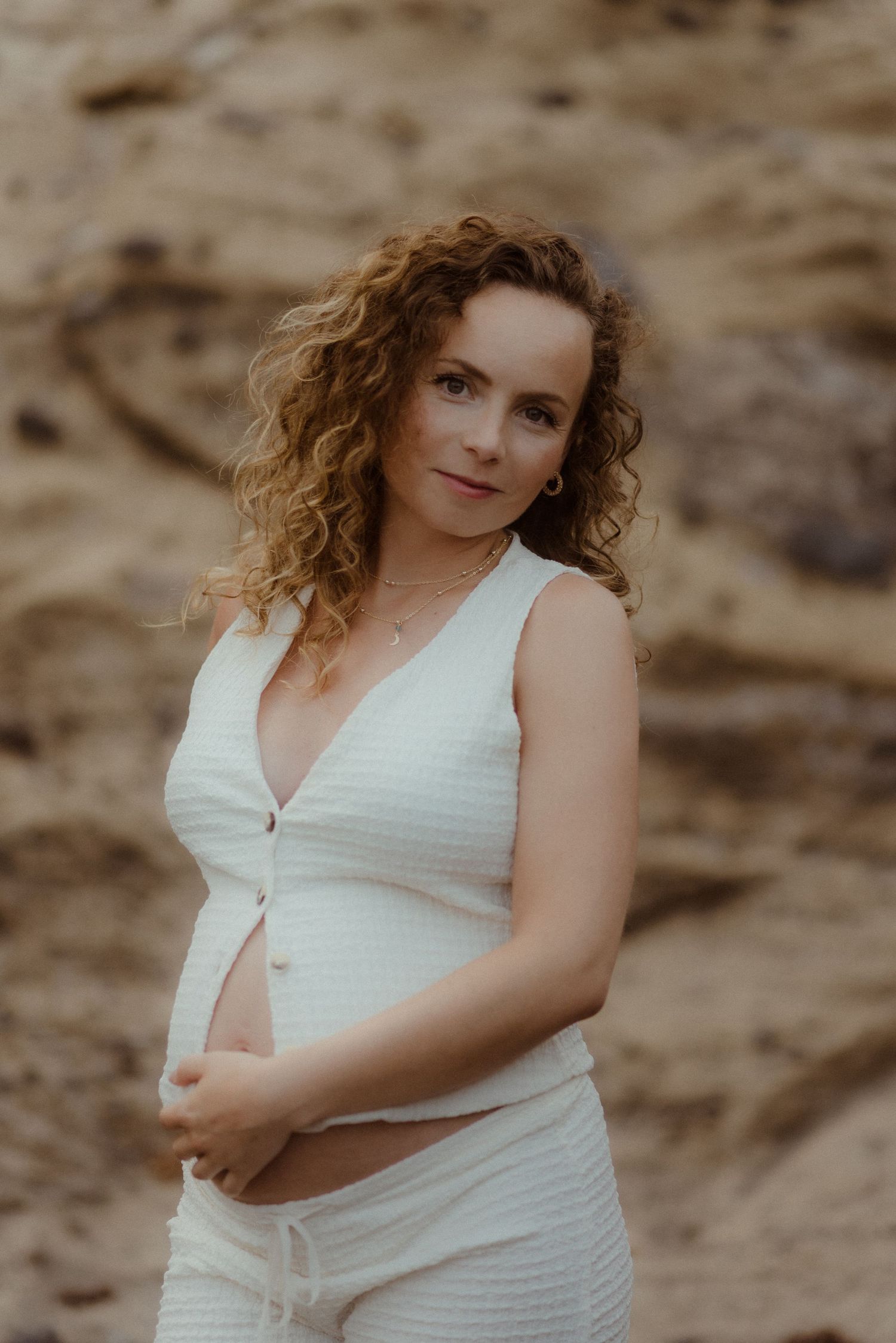 Expectant person in a white textured outfit poses outdoors against a sandy backdrop in warm natural lighting in El Médano, Tenerife.