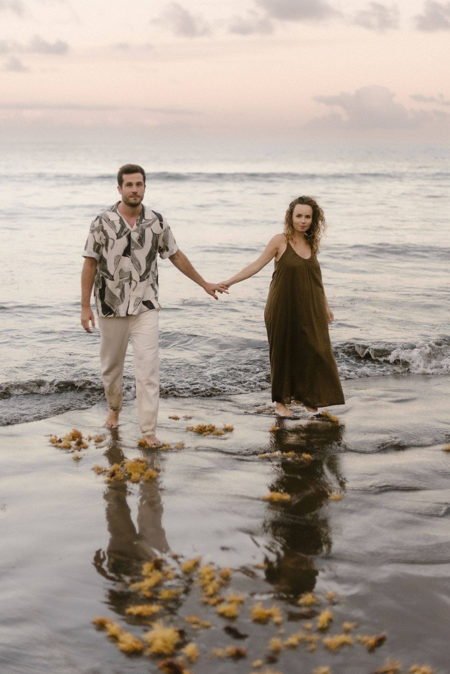 Two people standing in shallow ocean water at sunset holding hands with reflections in wet sand and beach waves in El Médano, Tenerife.