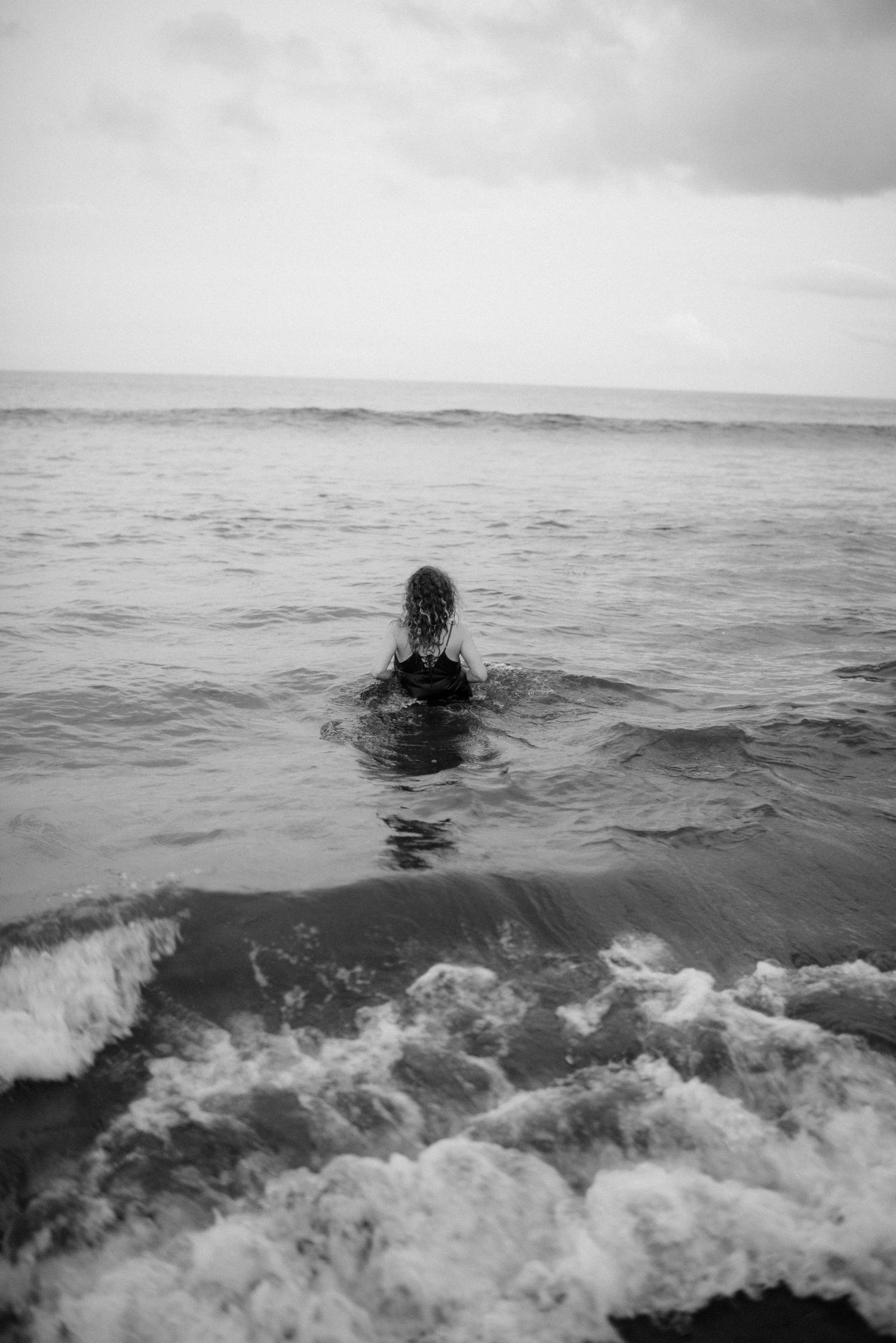 Black and white photo of a pregnant woman swimming in rough ocean waves against a cloudy sky in El Médano, Tenerife.