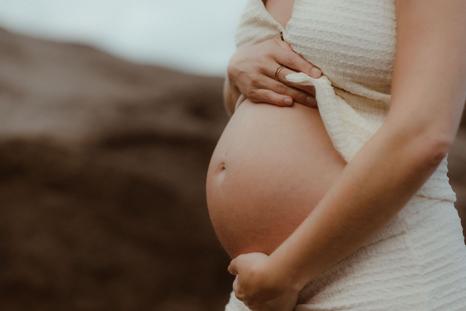 A pregnant woman in a white dress cradles her belly against a soft blurred background in El Médano, Tenerife.