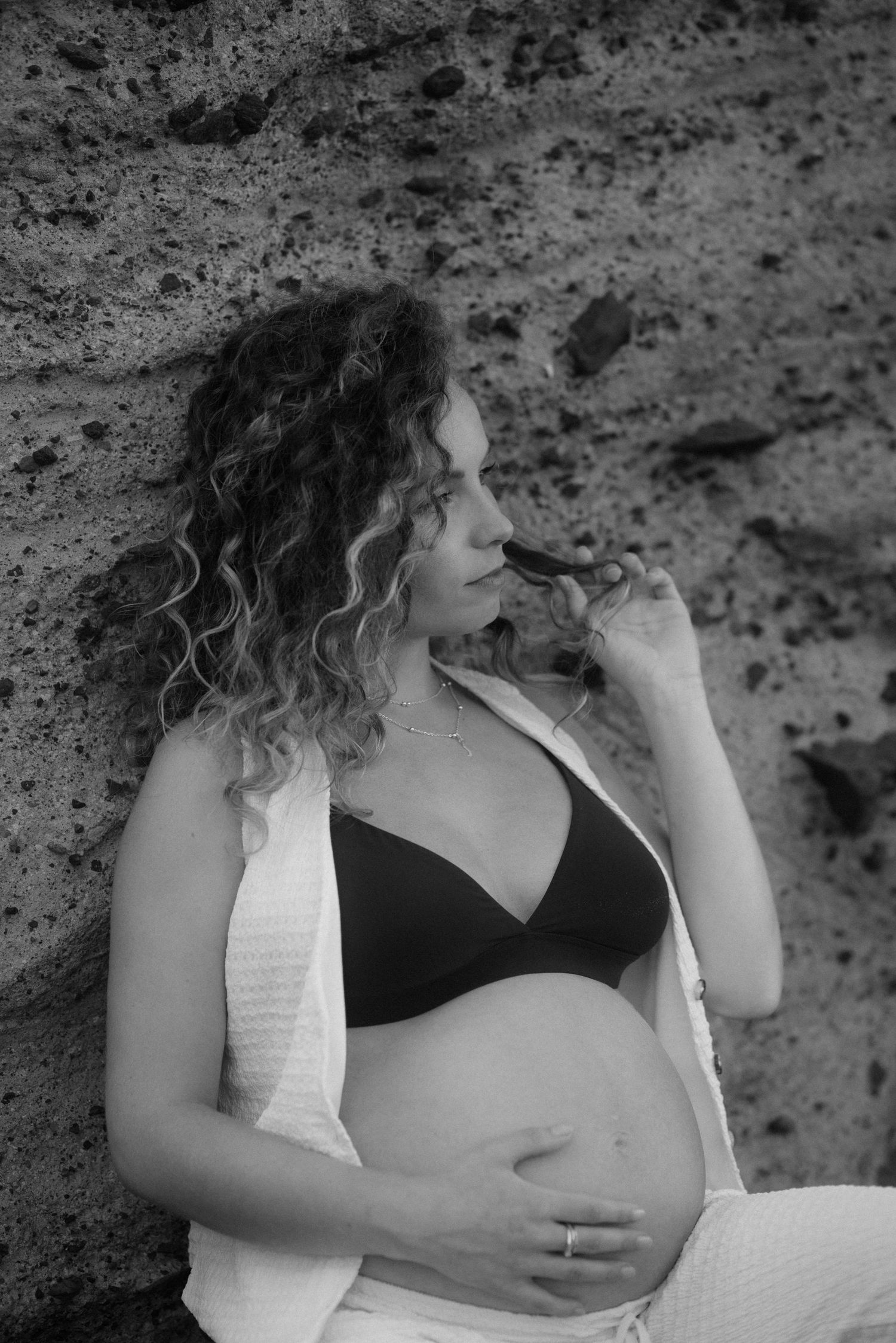 A pregnant woman in a black bikini top and white cardigan poses against a rocky background in black and white in El Médano, Tenerife.
