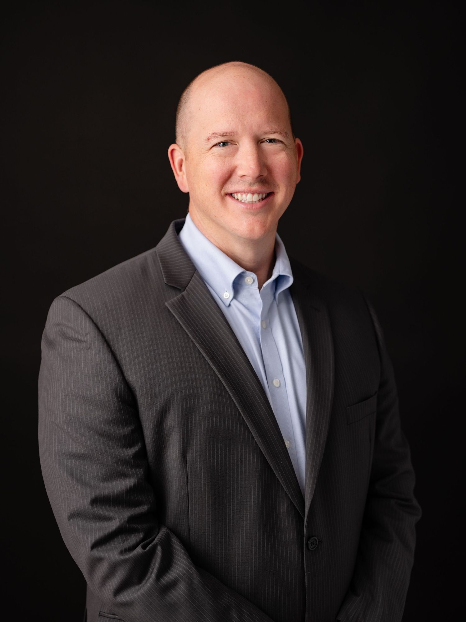 Professional headshot photo on black background showing person in pinstripe suit and light blue dress shirt.