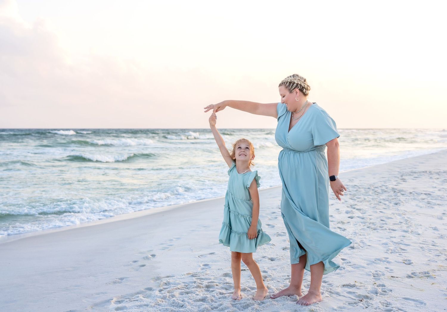 Two people in matching blue dresses dance together on a white sand beach at sunset as waves roll in along the shore.