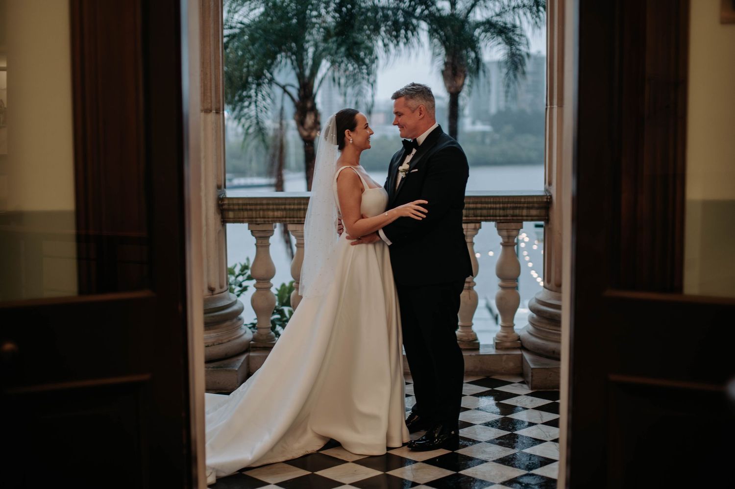A romantic wedding scene on a balcony with a checkered floor and palm trees visible in the background.