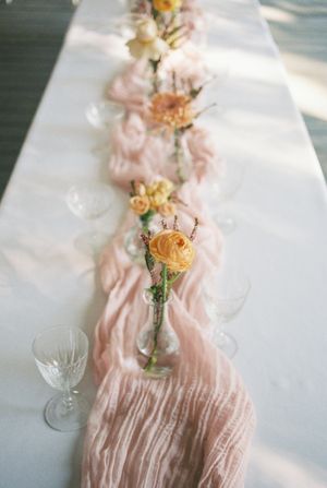 Delicate pink fabric table runner with yellow roses in glass vases creating an elegant wedding table setting.