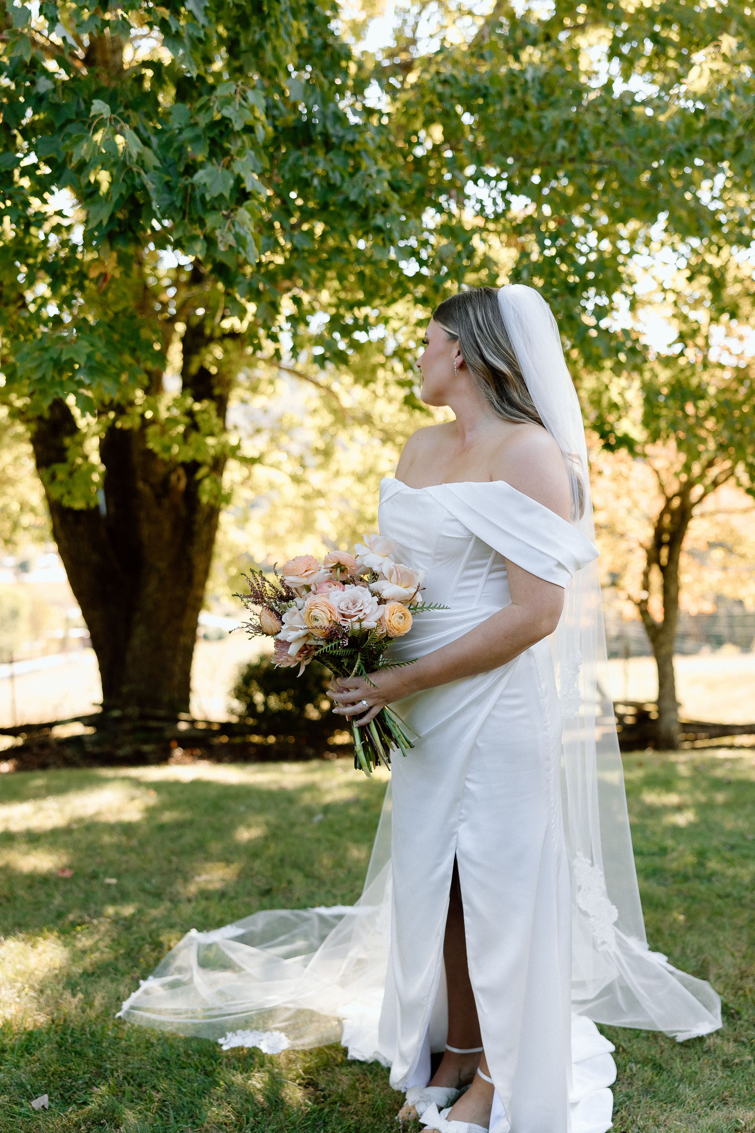 Person in an elegant off-shoulder wedding dress holding a bouquet in a sunny outdoor setting.