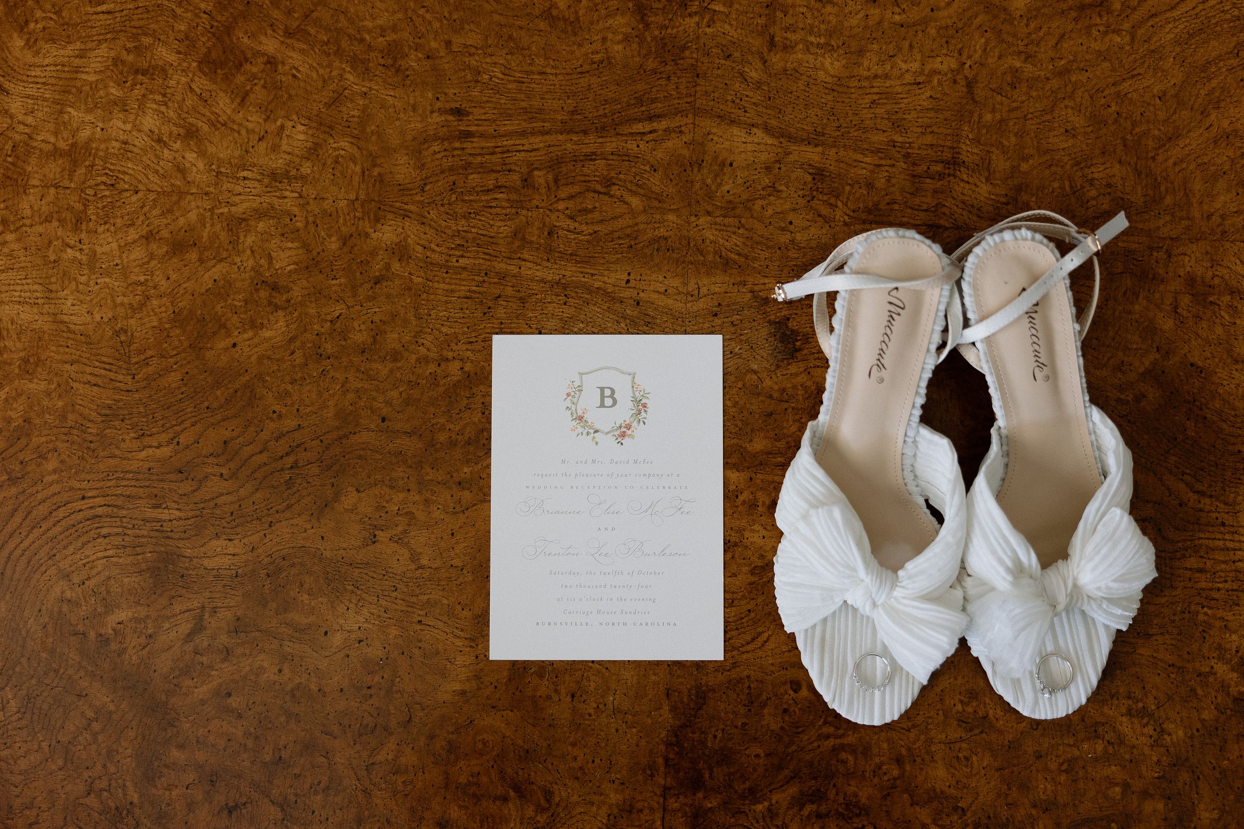 Elegant white bridal shoes and wedding invitation laid on wooden surface.