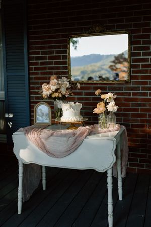 Elegant vintage white table with wedding cake and floral arrangements against brick wall with mountain view window.
