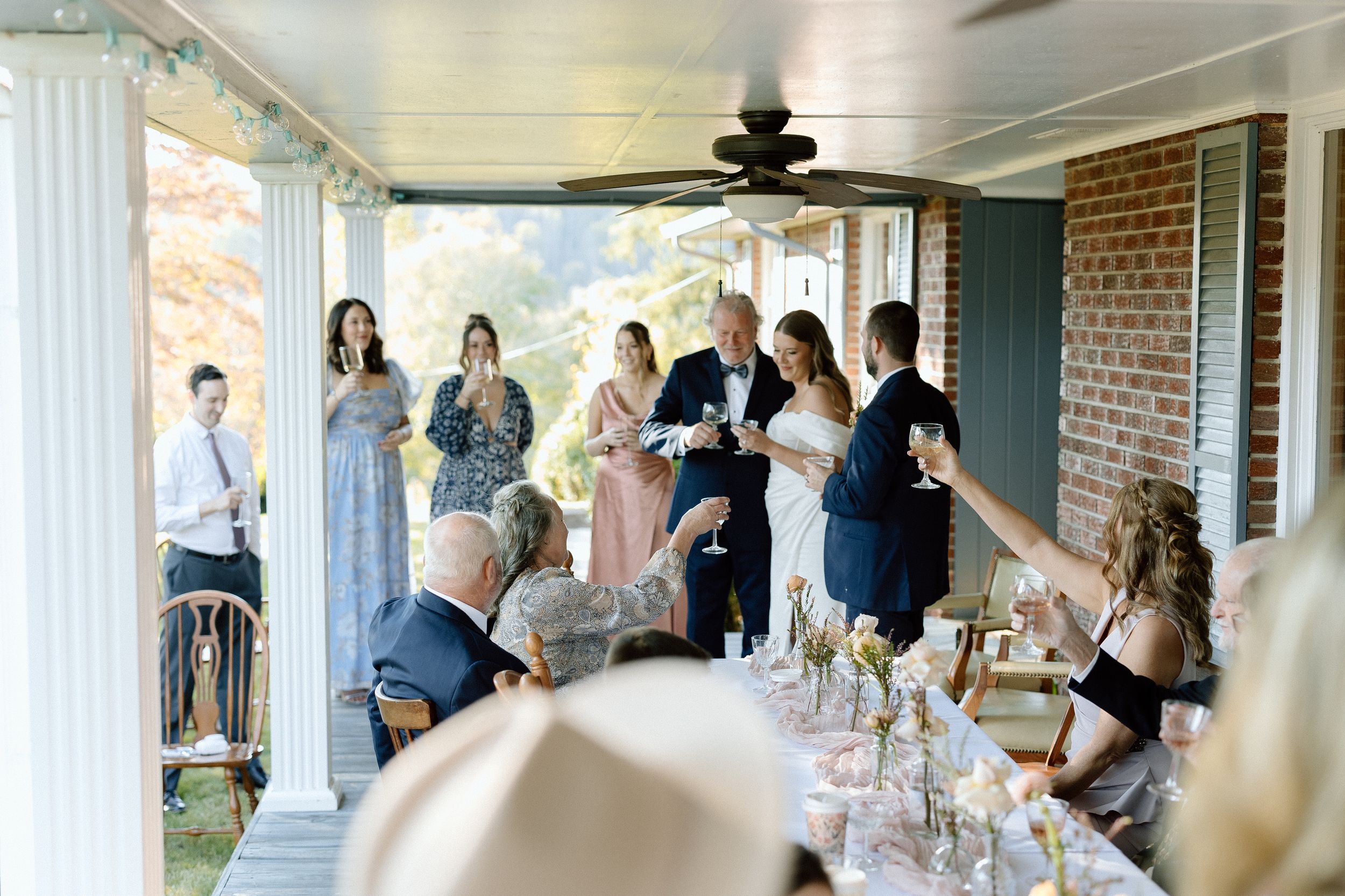 People gathered on a covered porch for a celebration with dining tables and brick wall backdrop.