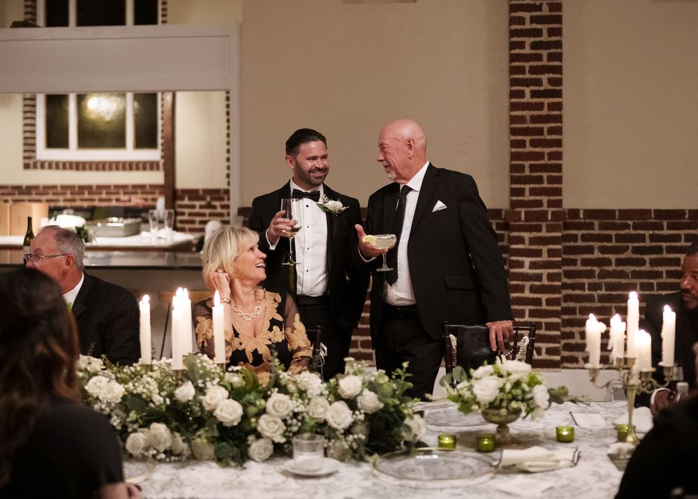 Elegant formal dinner party with white floral centerpieces and glowing candles on tables in a brick-walled room.