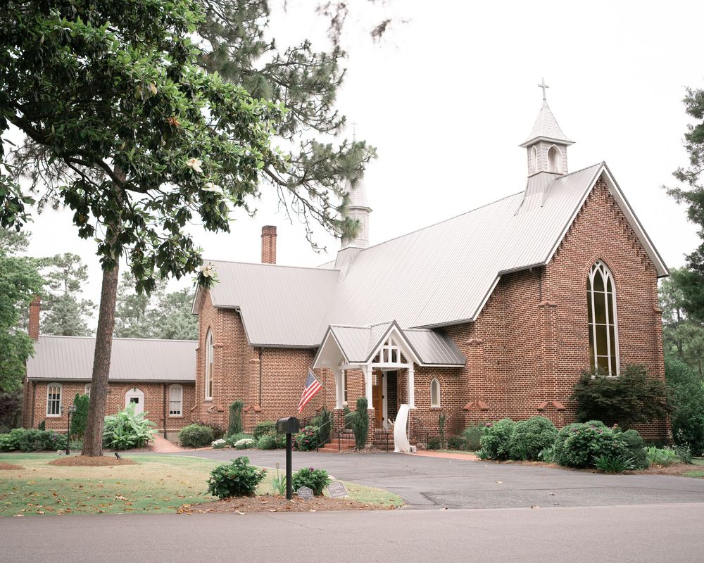 Red brick church building with white steeple set against evergreen trees and a paved parking area.