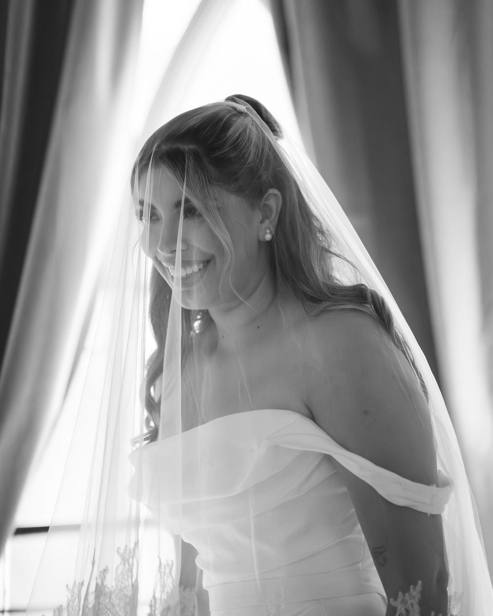 A bride in a flowing white wedding gown poses elegantly behind sheer curtains in black and white photography.