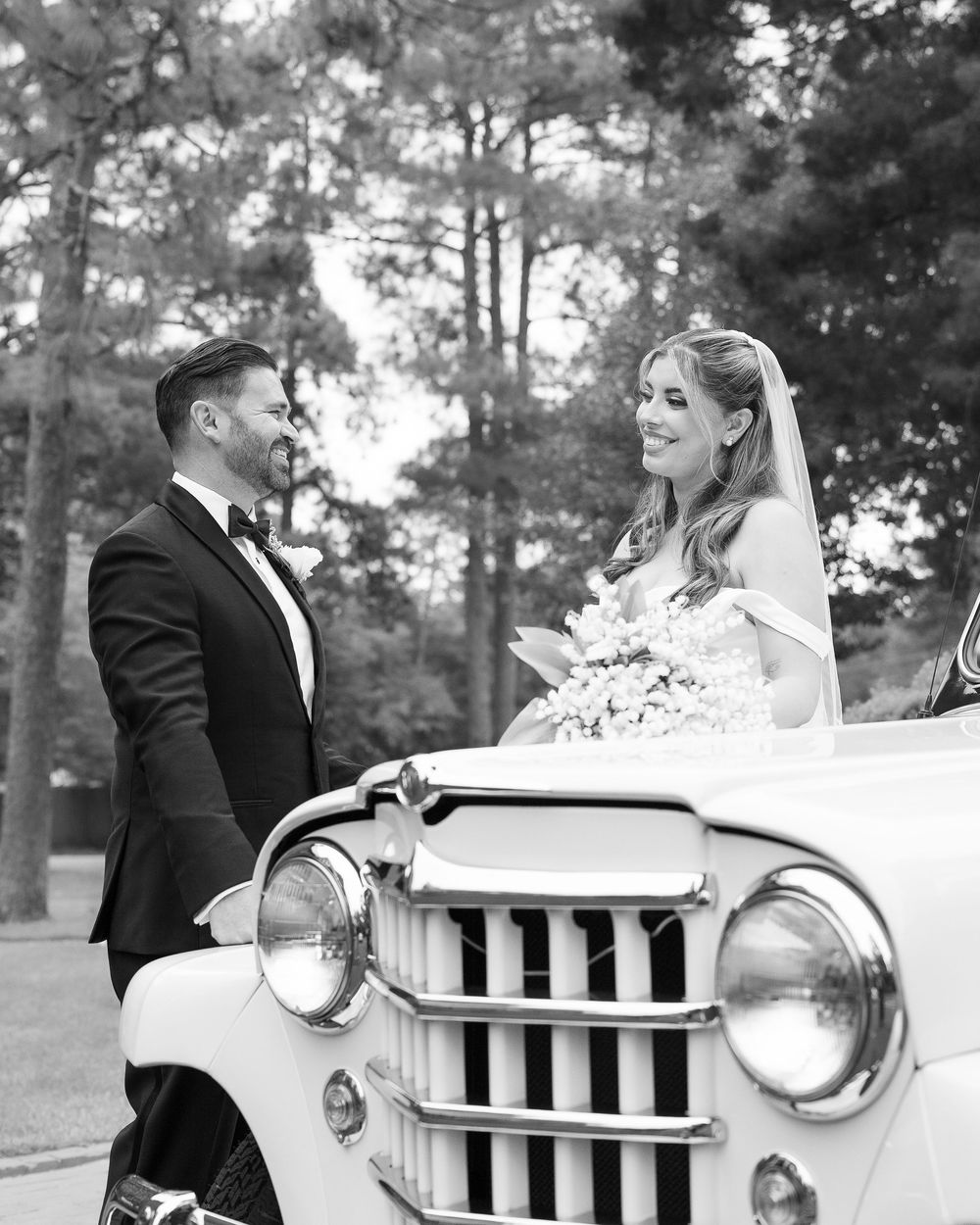 A bride in white dress and groom in tuxedo pose with vintage white Jeep surrounded by pine trees at outdoor wedding.