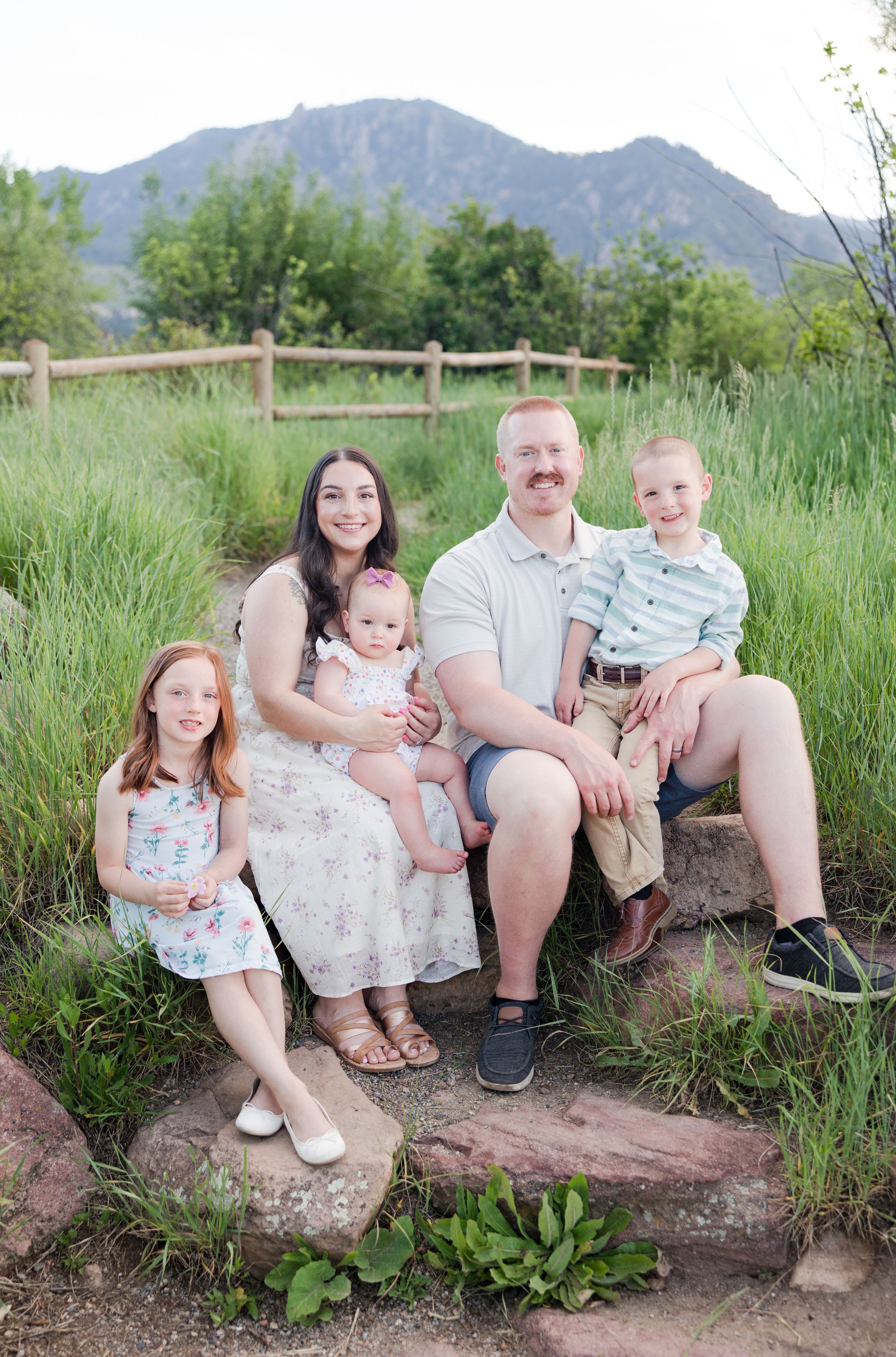 A loving family sits together on rocks with a scenic mountain backdrop in a grassy outdoor setting near a wooden fence.