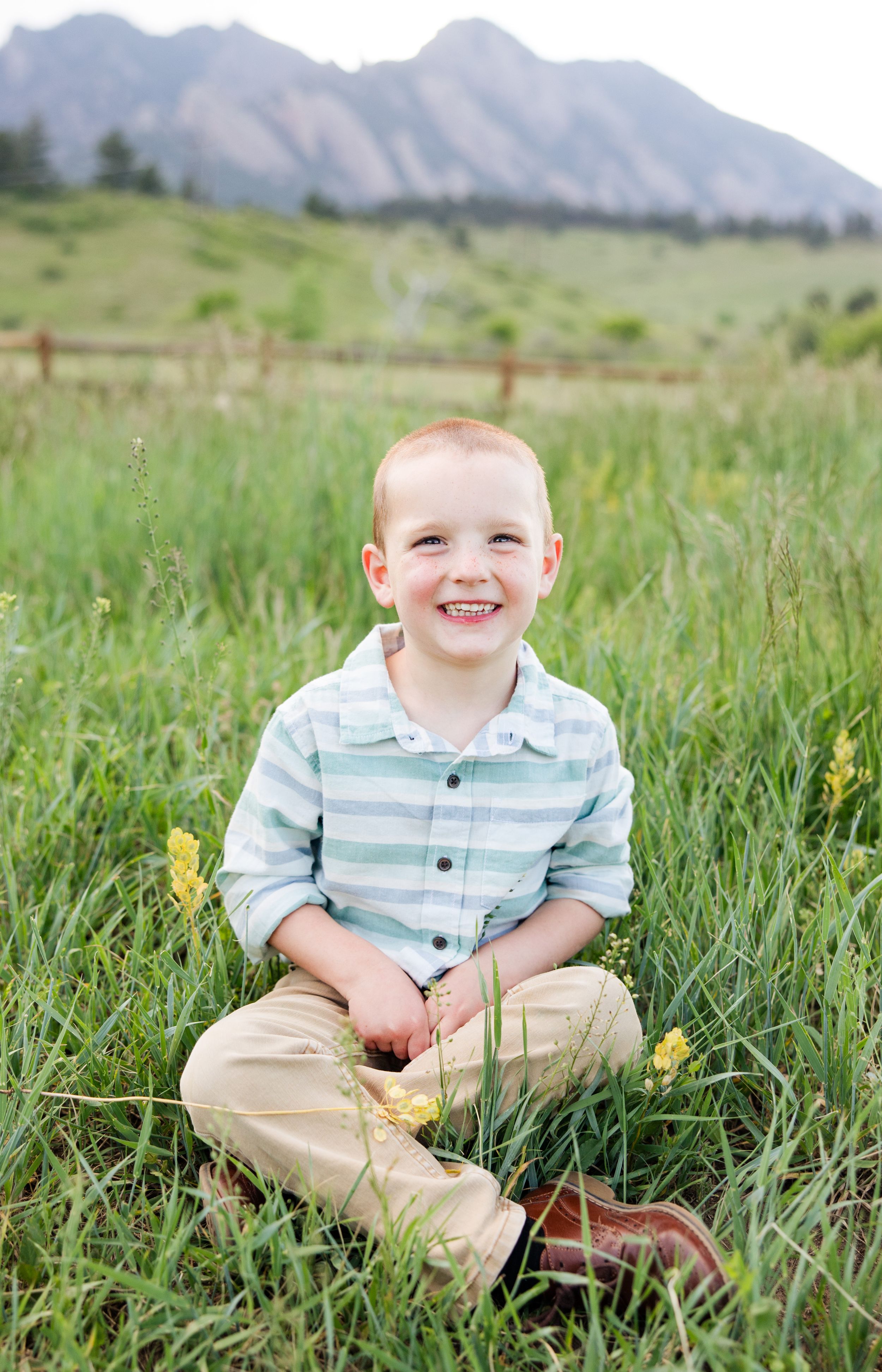 A smiling young child sits in a green meadow with mountain peaks visible in the background on a sunny day.