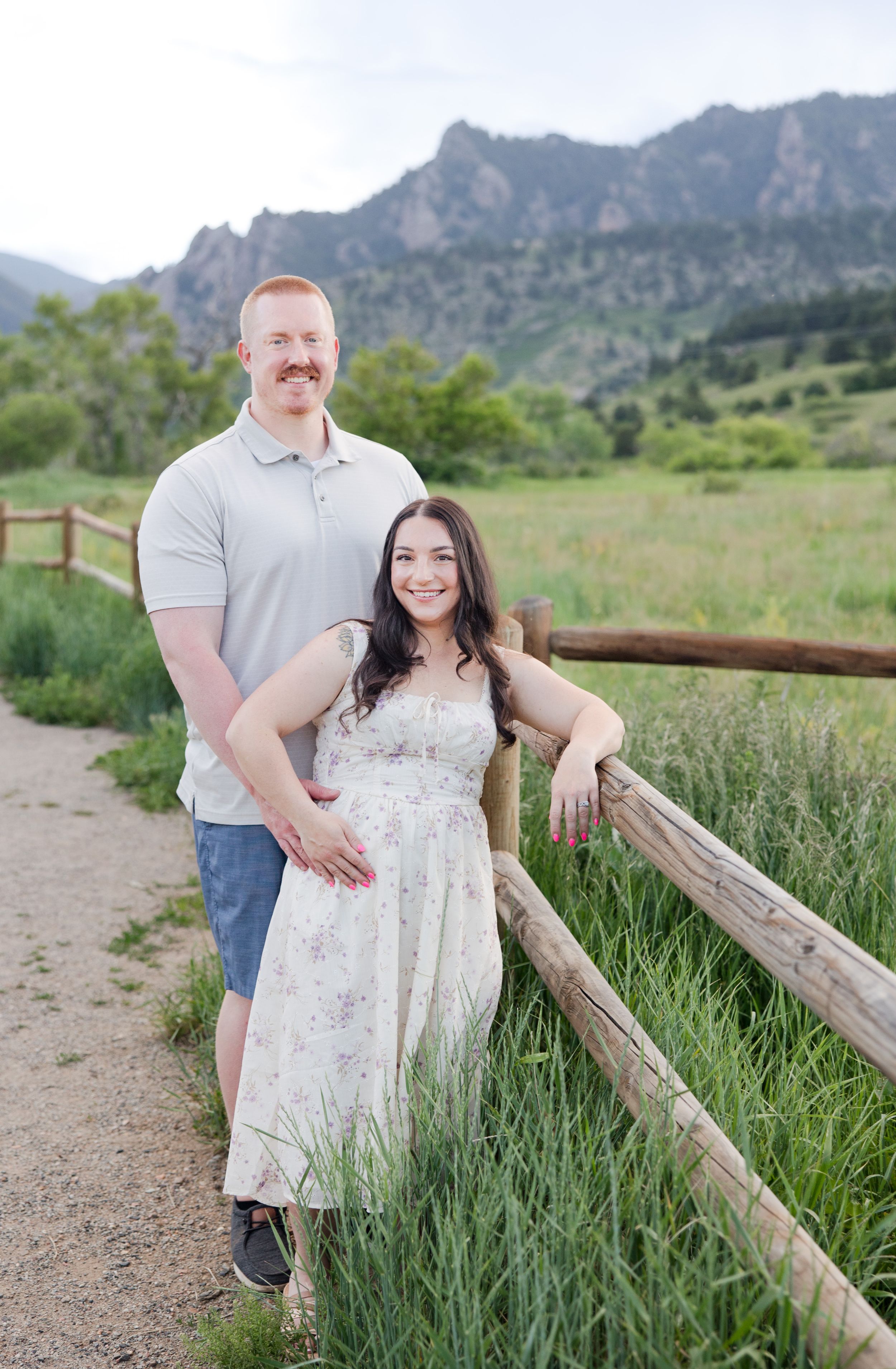 A couple poses together along a wooden fence with scenic mountain views in the background at sunset.