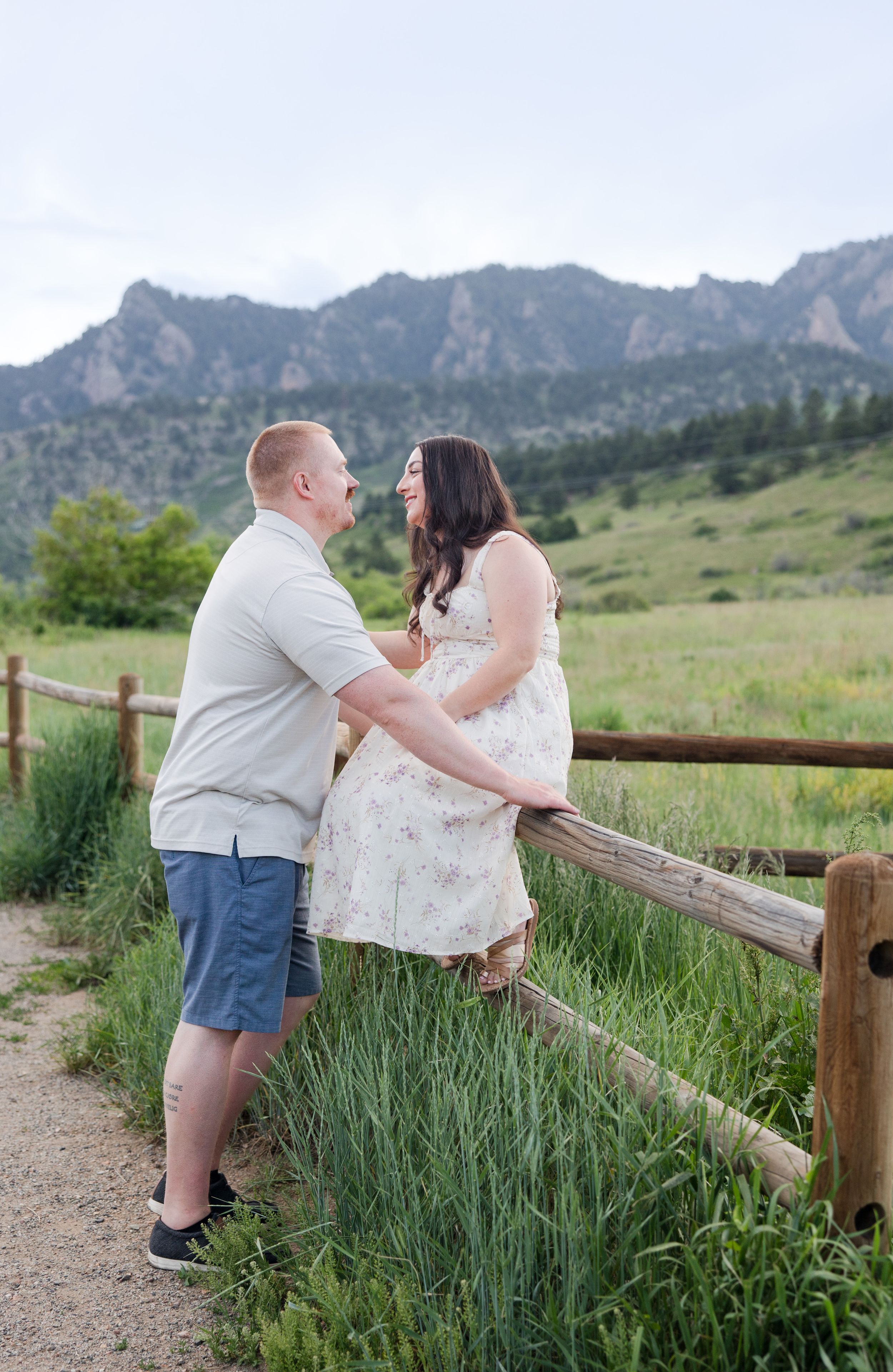 A couple shares a tender moment by a wooden fence with mountains in the background at sunset.