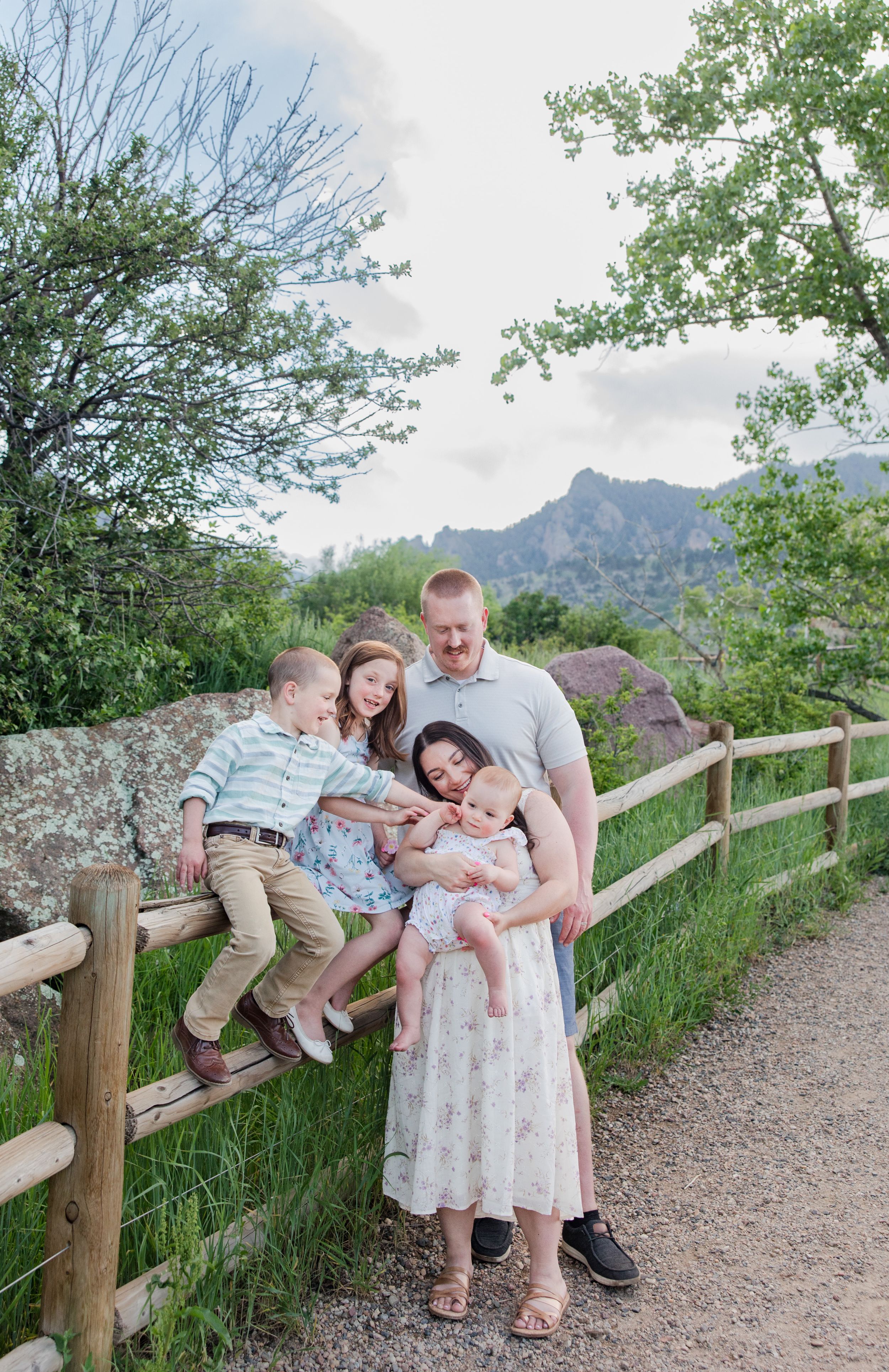 A group poses together by a wooden fence on a mountain trail with rocky peaks and green trees in the background.